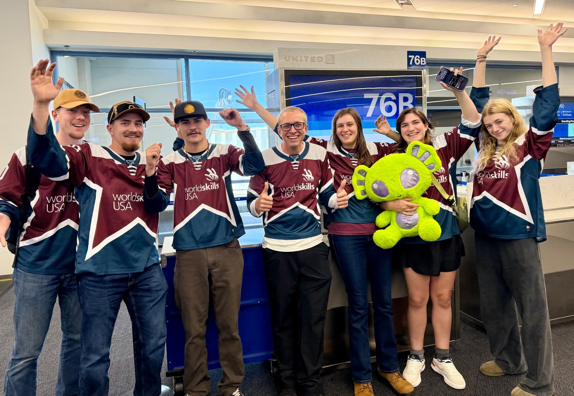 Seven WorldSkills USA competitors in team jerseys celebrate at an airport departure gate, with one holding a green mascot.
