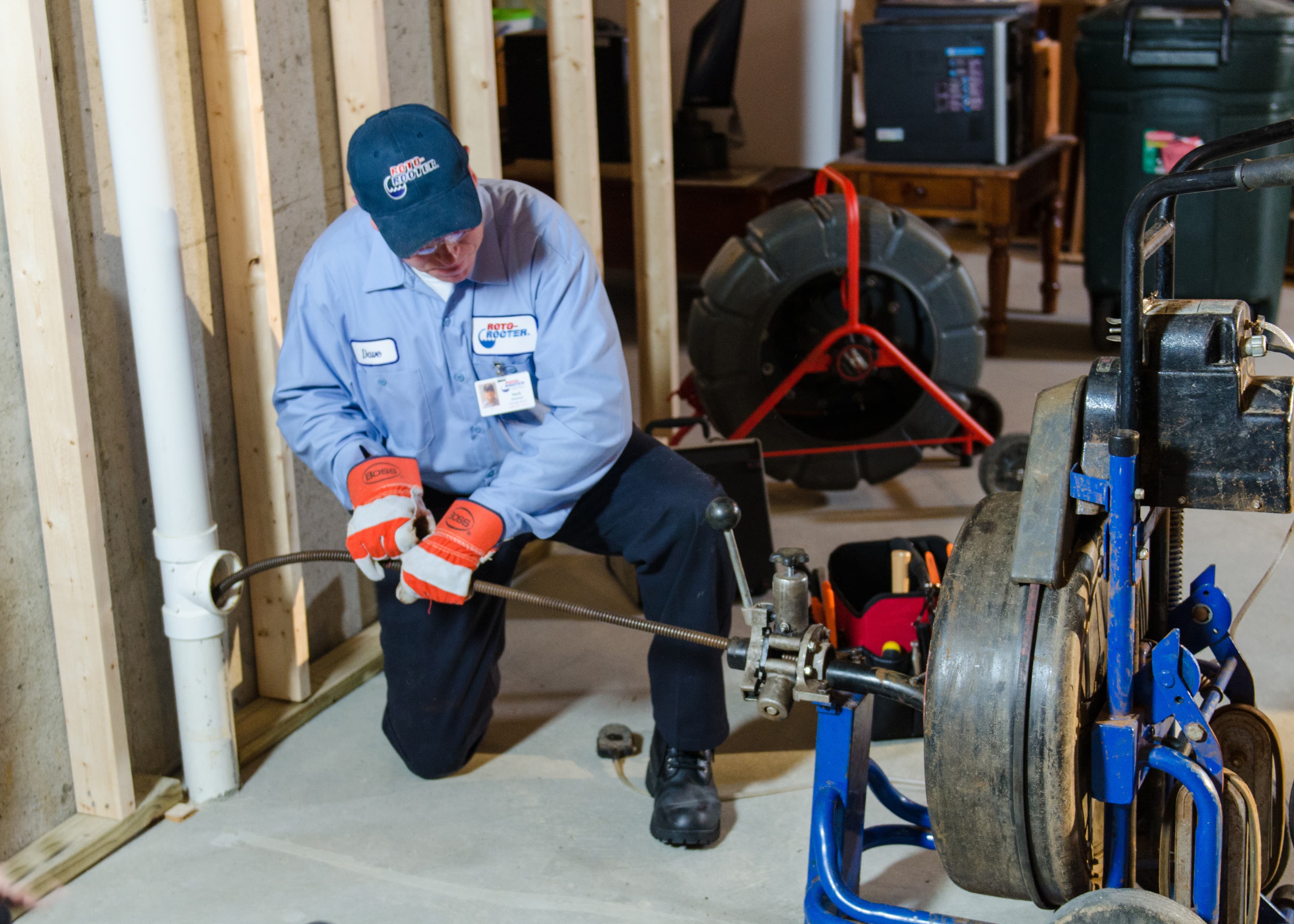 Roto-Rooter technician in a blue uniform operating a sewer-cleaning cable machine beside a basement drainpipe.