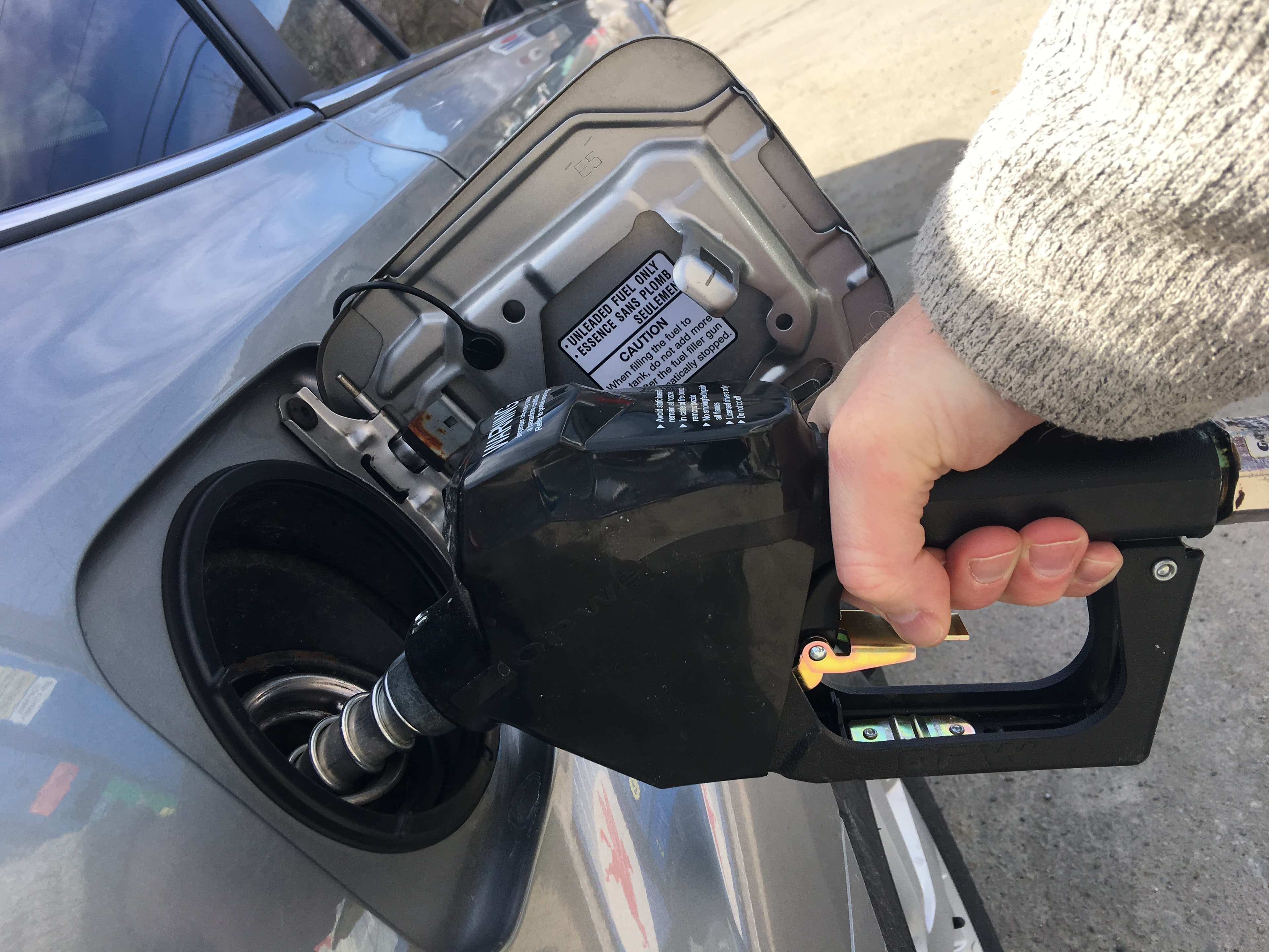 Close-up real photograph of a driver holding a gasoline nozzle while refueling a vehicle in the United States.