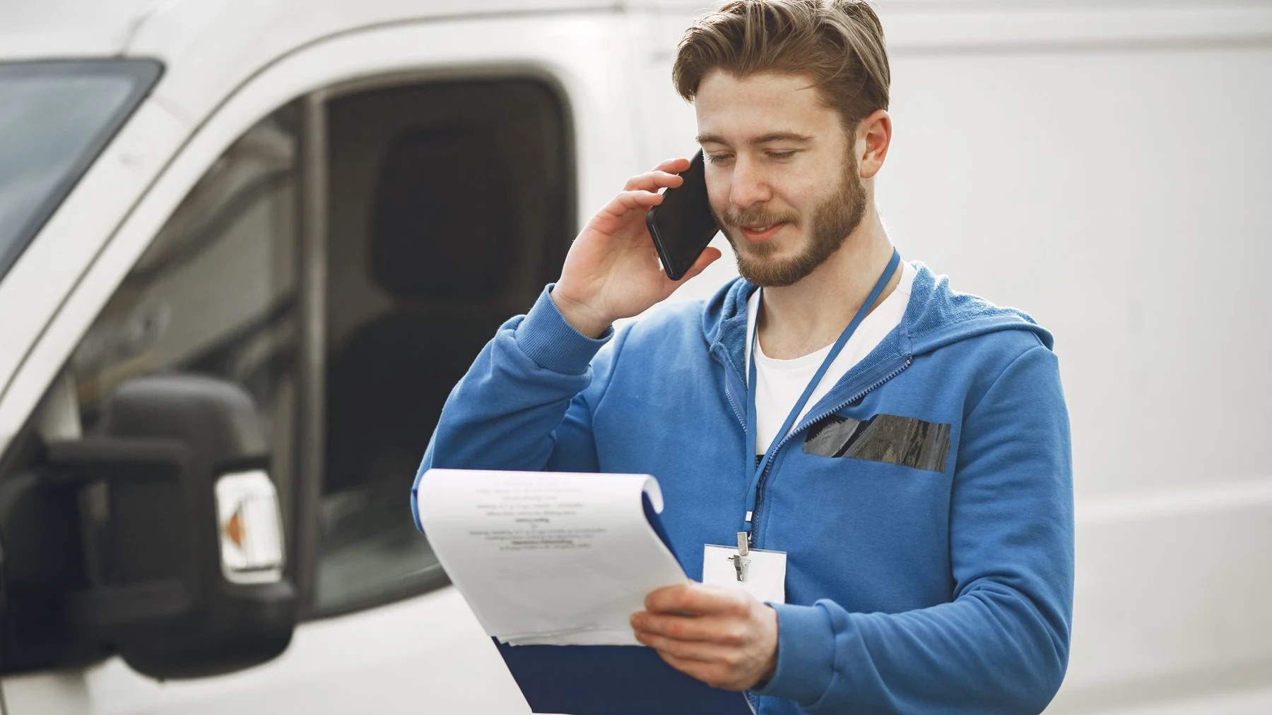 A worker beside a white van takes a mobile phone call while holding a clipboard.